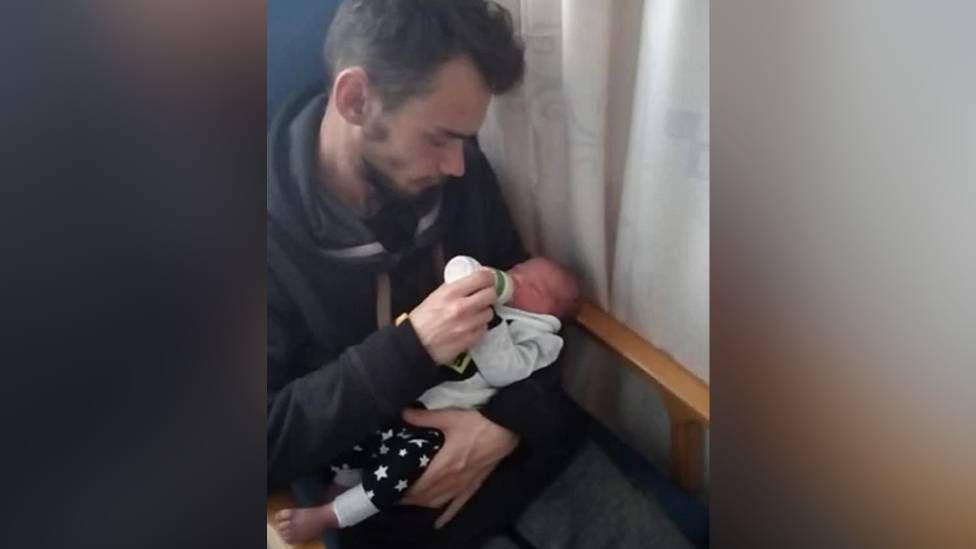 Will is seen feeding a bottle to newborn baby Ethan. He is sat down in a chair with a wooden frame with a white curtain on the right hand side.