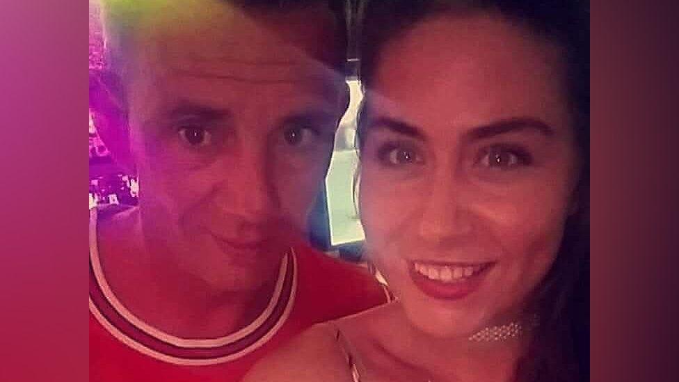 Gary Burns with short brown hair and brown eyes wearing a red t-shirt with his sister Danielle with long dark hair wearing a dress and a pearl choker in a bar in Turkey. They are both smiling.