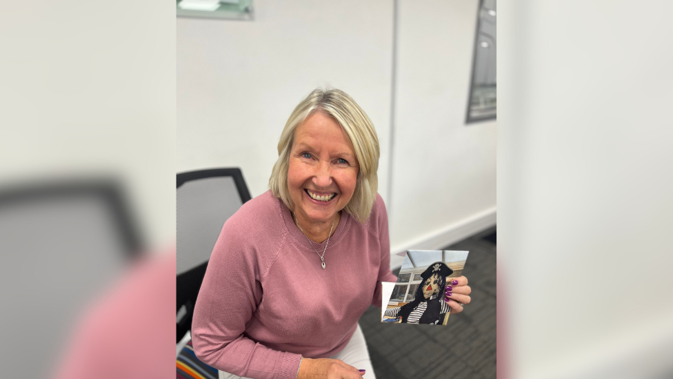 Ms Coull is sat smiling at the camera. She has blonde hair, wearing a bit jumper and holding a picture of her entry - a pirate scarecrow.