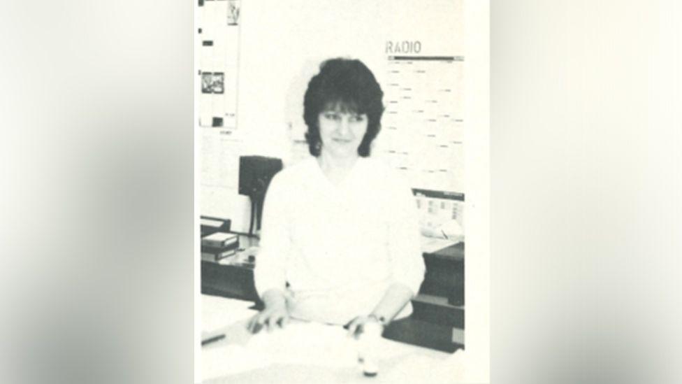 Pat pictured in black and white standing with her hands on a photocopier or desk and glancing slightly to one side. She wears a white blouse and has black curly hair in a bob.