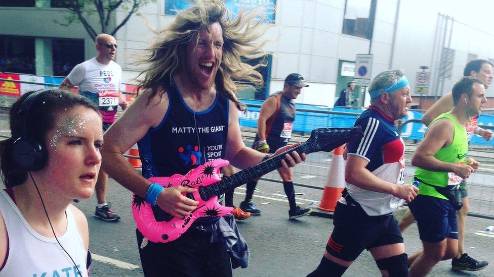 A marathon runner holds an inflatable pink electric guitar. He has long hair and a big smile.