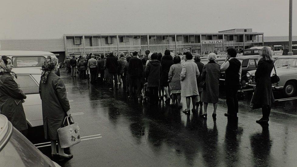 A black and white image of people queuing in a carpark in front of a Sainsbury's store in 1975