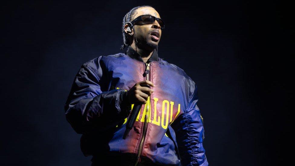 21 Savage performs his first ever London headline show at The O2 Arena on November 30, 2023 in London. The rapper has braided hair and wears sunglasses and a blue and red bomber jacket. He holds a microphone and is pictured on stage, which is dark behind him.