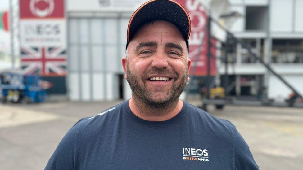 Man with orange edged baseball cap wearing a navy blue T-shirt he has stubble on his face and is smiling