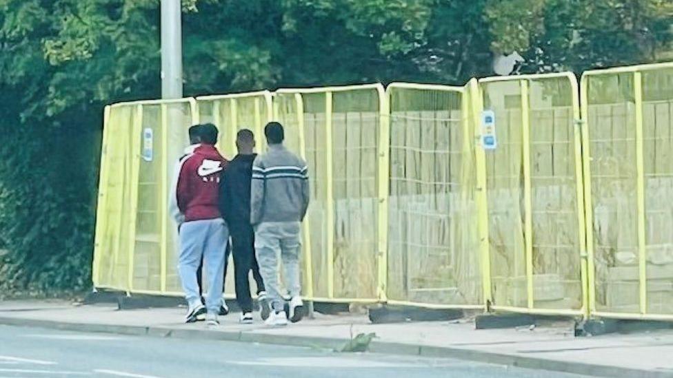 Four men, from behind, walking along a pavement, next to yellow fencing.