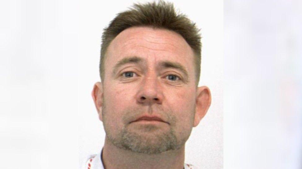 A police mugshot of a white man looking to the camera unsmiling. He has short brown hair and is unshaven