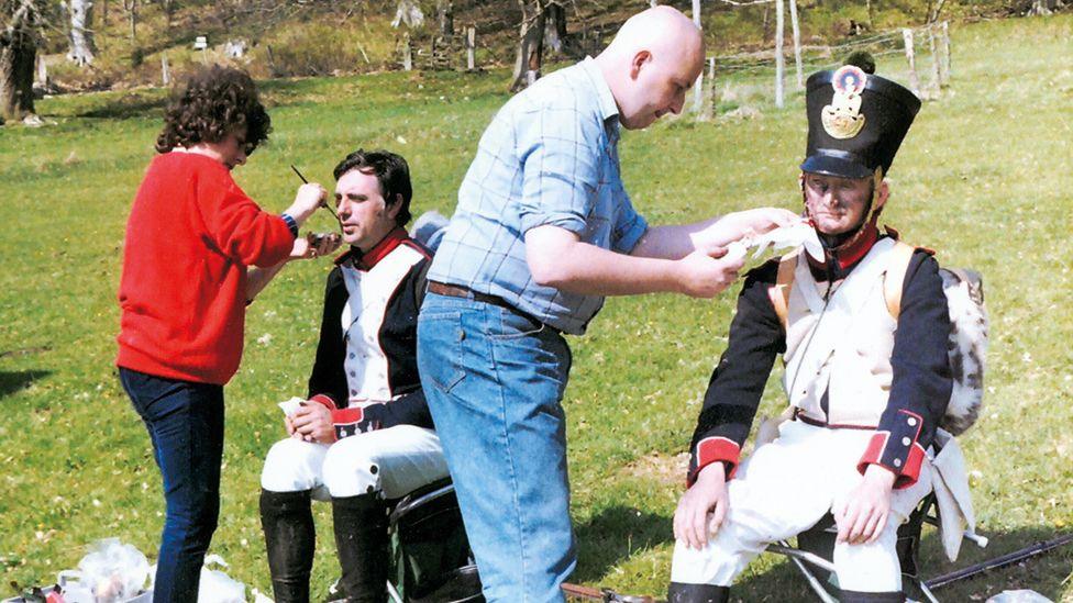 Two men, dressed in historical soldier outfits are having makeup applied to them. They are both sitting on chairs. A lady in a red jumper, with brown hair and jeans on, is applying makeup to one of the actors and a young Dave Myers is applying makeup to the other actor. He is dressed in a checked shirt and blue jeans. He has a look of concentration on his face.