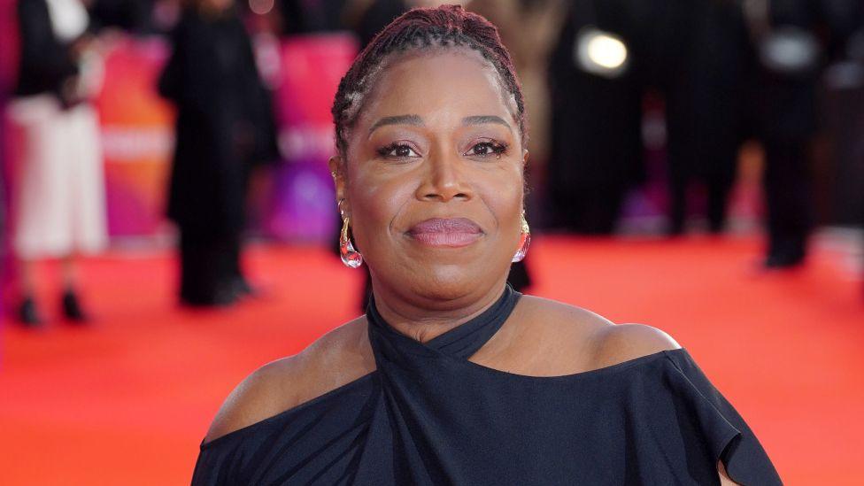 Michele Austin smiles at the camera. She's wearing a halterneck black dress and the red carpet can be seen in the background