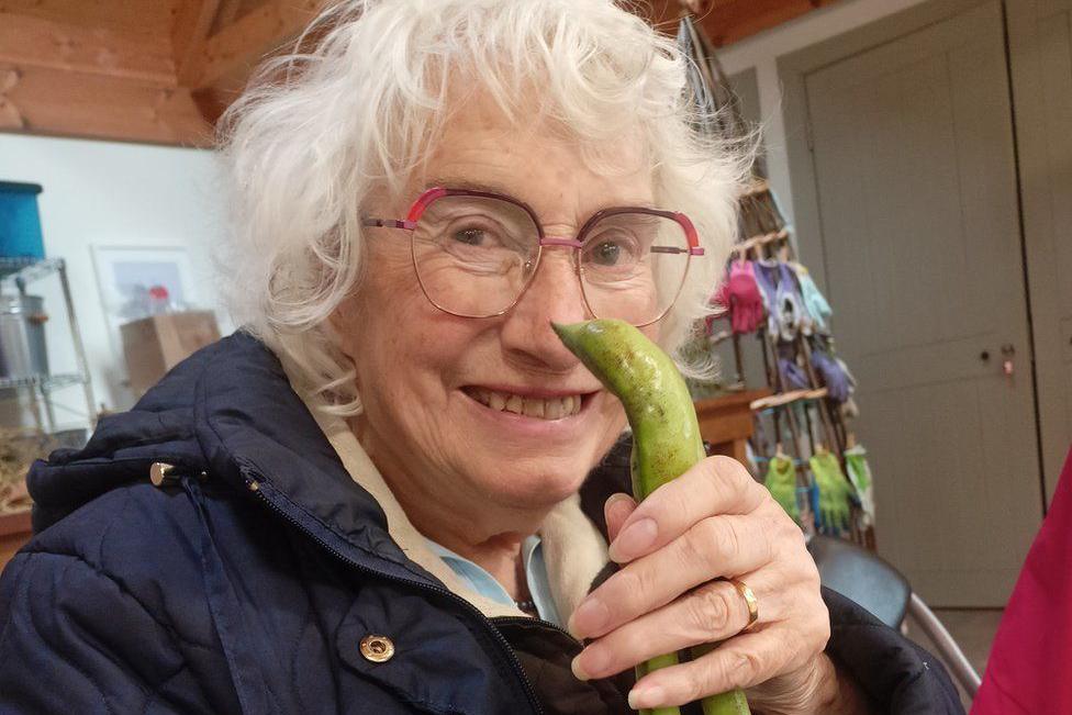 Maureen is holding up two large runner beans in front of her face and smiling. She has glasses, a blue padded coat and grey hair.