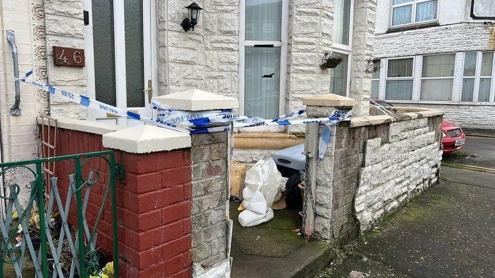 The small front yard of a terraced house, with piles of rubbish between the low front wall and the front of the house. Police tape has been placed across the gate posts. The brickwork of the house appears to have been covered in white "brick effect" cladding. Similar cladding on the front yard wall has come away exposing sections of the normal house bricks underneath.