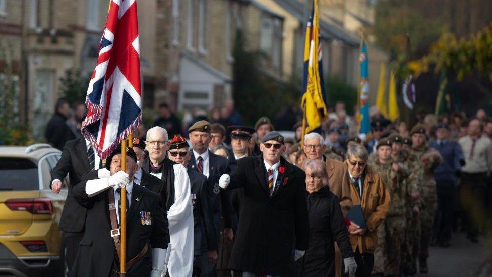 Parade led by the union flag carried by an RBL member with a priest alongside. Other standards are visible in the background