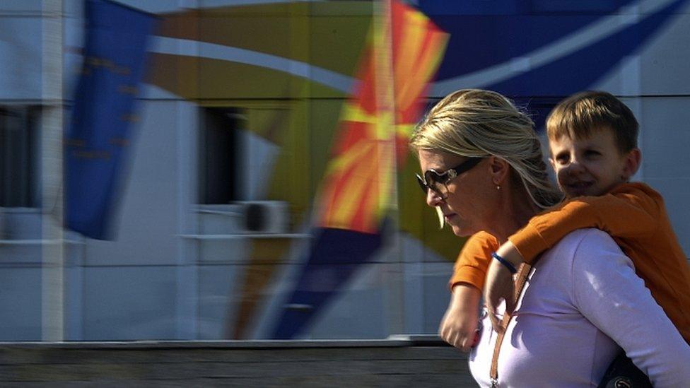 A woman carrying her son on her back walks in front of North Macedonian and EU flags in Skopje, North Macedonia, 18 October 2019