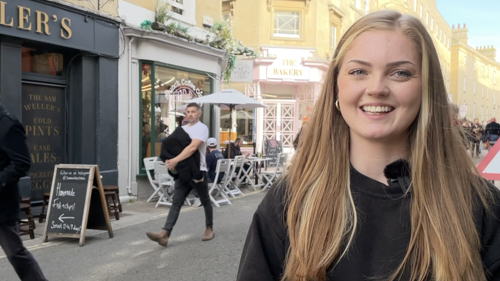 Olivia has long blonde hair. She is standing in the street. In the background you can see cafe tables and shoppers. She's wearing a black jumper. She is in the right of the frame.