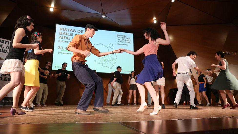 Groups dressed in early 20th century clothing twirl and step on a stage, in front of the voting results displayed on a large screen on the wall nearby