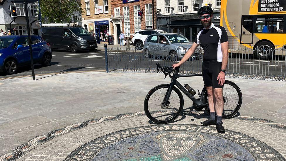 Richard Slade is standing in a town centre in front of a mosaic on the pavement. He is holding a black bicycle. He is wearing a tight-fitting blue and white cycling top and shorts, a bicycle helmet and has sunglasses on. He has a greying beard and moustache.