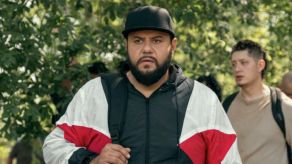 A still from the show, when Mo, wearing a black, white and red jacket, returns to his family's home. There are green leaves behind him and people walking behind him.