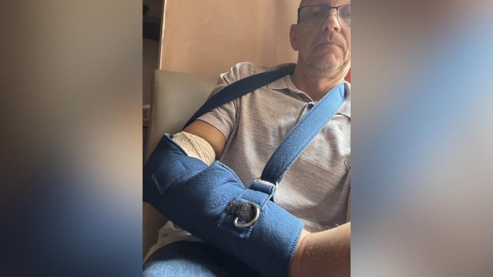 A middle-aged man sits in a chair with his arm bandaged and in a blue sling.