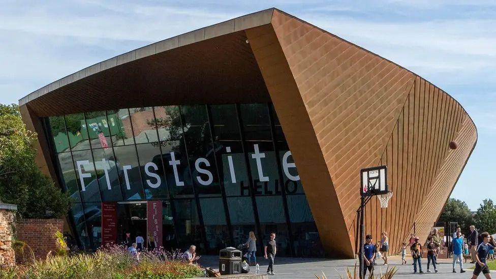 A modern wood-panelled museum building with small crowds of people walking around outside. There are blue skies above.