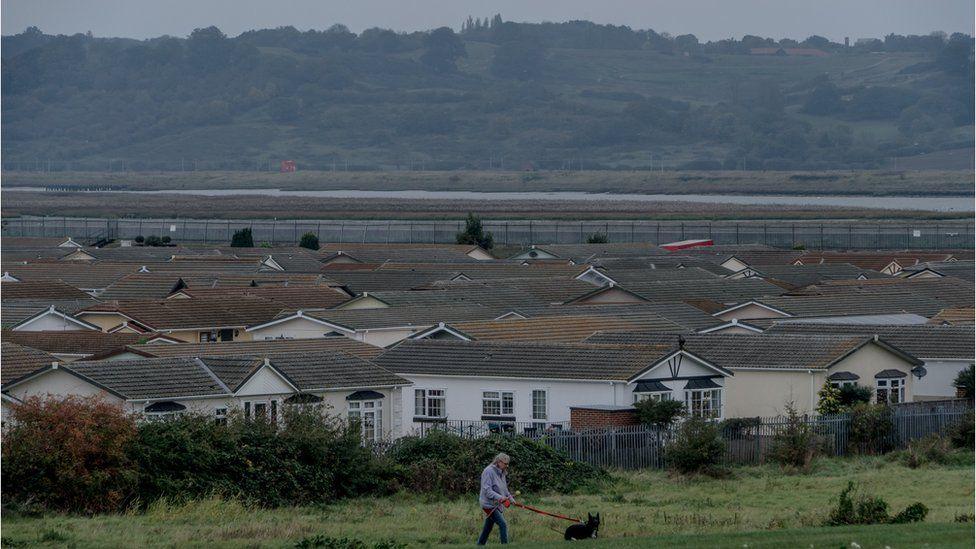 A wide shot of a series of white houses with grey roofs. In front of the houses there is green land and a man wearing a purple coat walking a black dog.