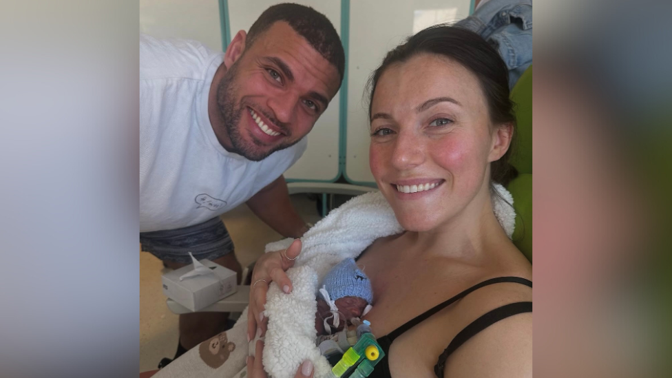 Zack George leaning down next to his wife Samantha who is in a hospital bed with their premature son Leo on her chest.