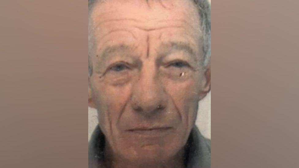 Brian Fletcher stares forward in a dated photo released by police. He is wearing a grey shirt and has grey hair with a scar below his eye.