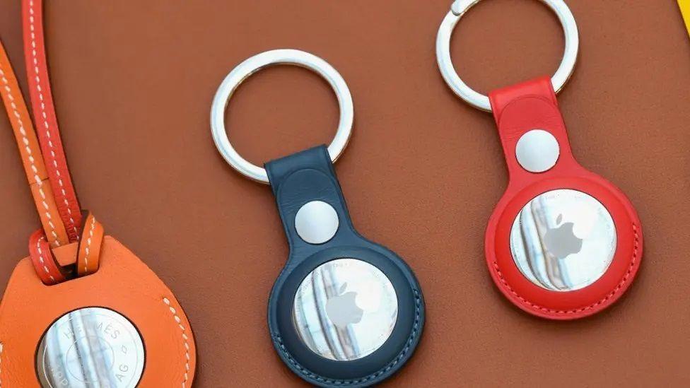 Three AirTags on different coloured keyrings. One is orange, another black and the third is red. Each has a large metal disc with the Apple logo in the centre of the fob and a smaller metal disc between the larger one and the ring.