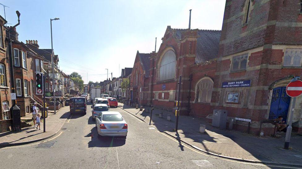 Bury Park Road seen from a crossroads. On the right is a United Reformed Church large brick building, with terraced houses further down. A line of cars and vans are parked on the left of the road, and a line of vehicles are queuing.
