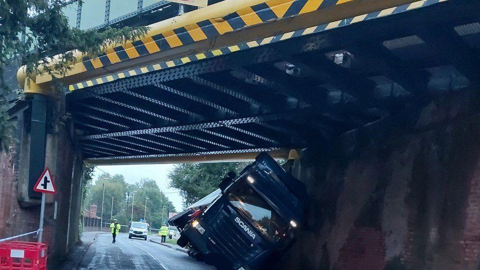 Underneath of metal and brick road bridge with lorry wedged underneath. The cab and trailer are leaning over on to the wall of the bridge