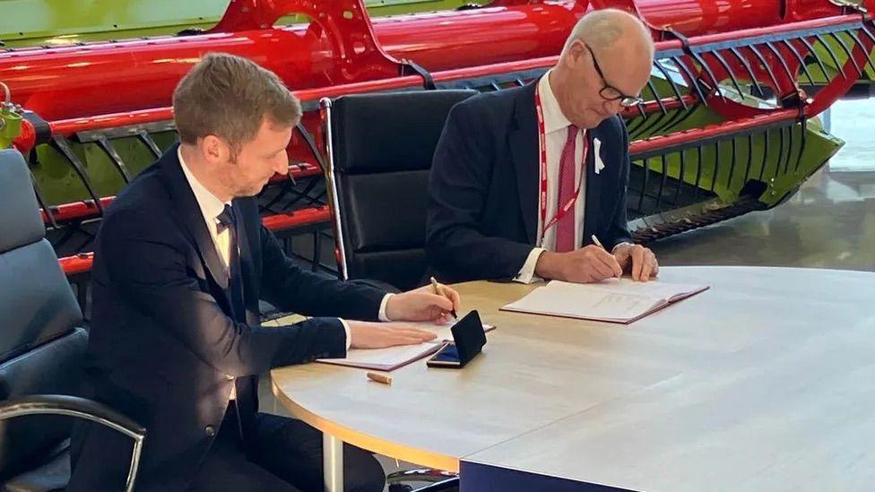 Former Local Government Minister Lee Rowley and Suffolk County Council leader Matthew Hicks, both in suits, sitting at a desk signing devolution papers in 2022. They are both holding pens and looking down at two books.