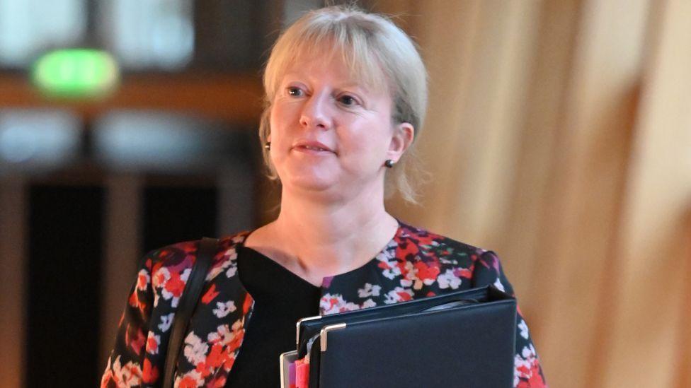 Shona Robison, who has blonde hair, looks to her right while walking towards the camera. She is carrying a black folder held against her chest. She is wearing a red and black floral top.
