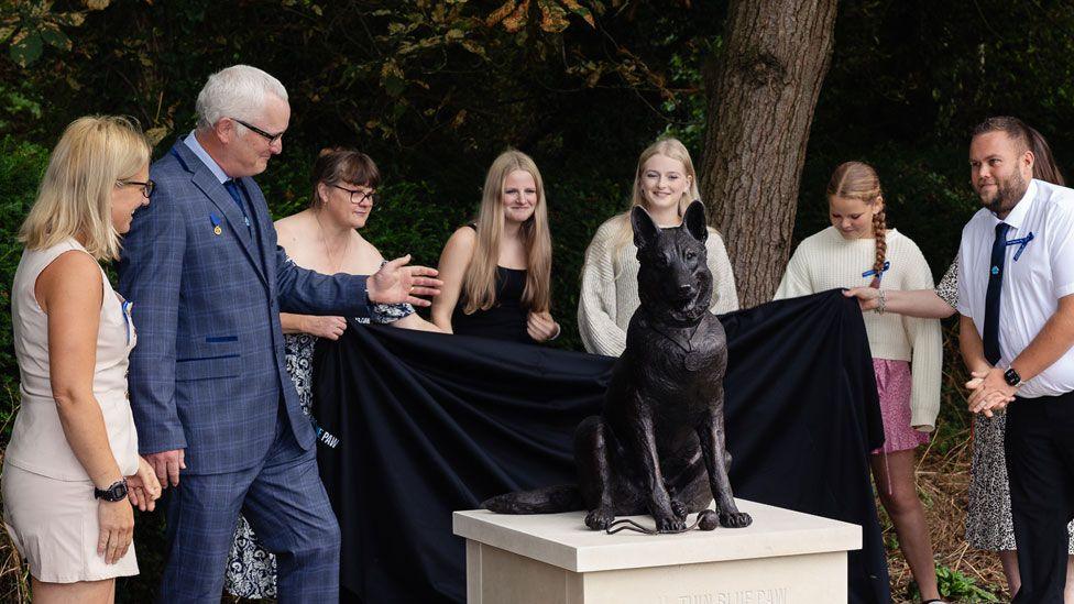 A group of seven people gathered around a bronze statue of police dog Finn. The statue shows Finn sitting up right with his ears pricked and his head tilted to the left. In front of him is ball. The statue is resting on a stone plinth. Five women - one hidden by a man - have just taken a black cloth off the statue.