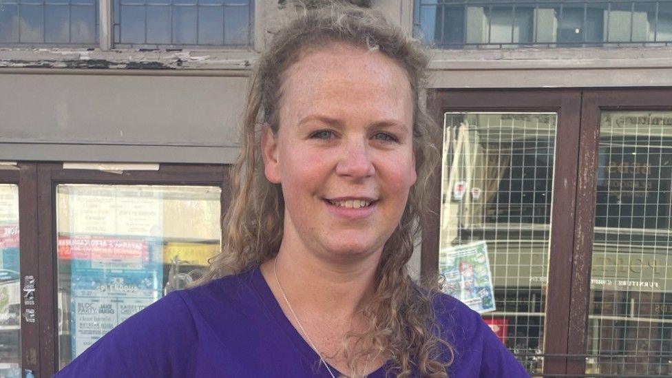 Anna Foster, who has long fair hair, is wearing a purple top and is standing in a street in front of windows with dark brown wooden frames.