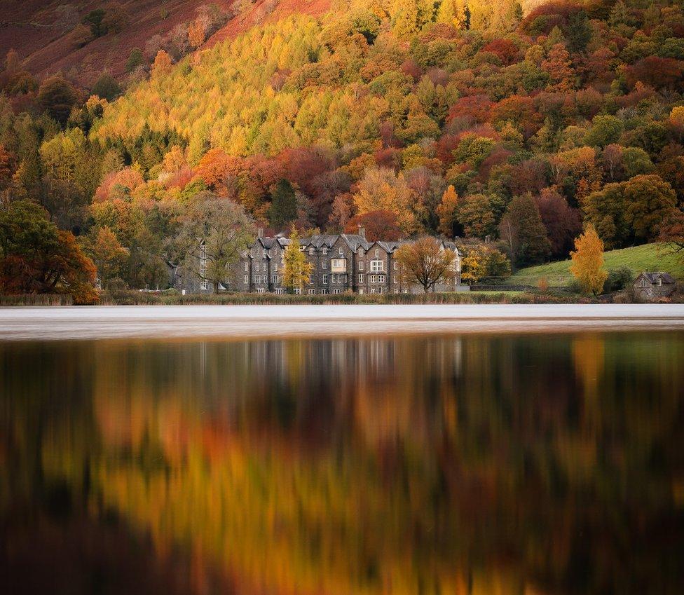 A large stone hotel surrounded by red, orange and green trees reflected in a lake