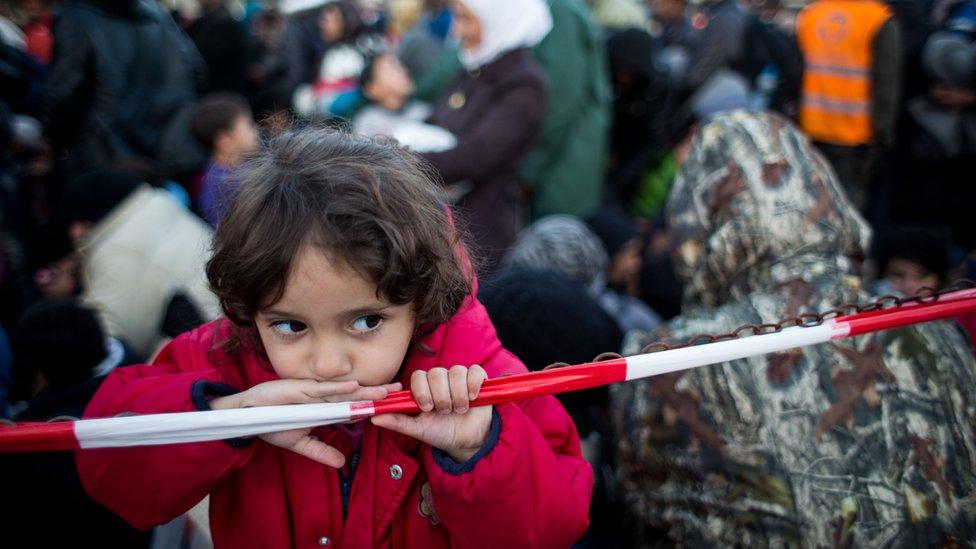 A girl plays with a chain barrier, with the background packed with refugees and migrants in this 2015 photo from Spielfeld