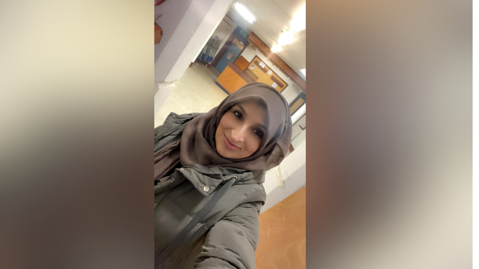 A young woman in a brown hijab and winter jacket smiles and poses for a selfie