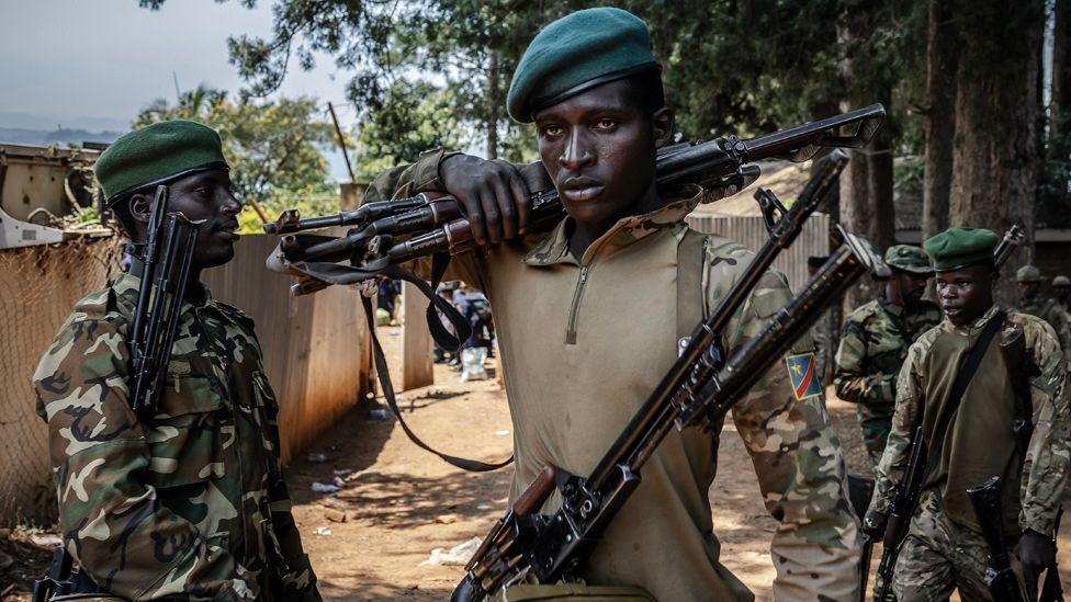 A member of the M23 movement in military fatigues carries weapons over his shoulder. Another fighter is to his right and he is holding weapons under his arm.