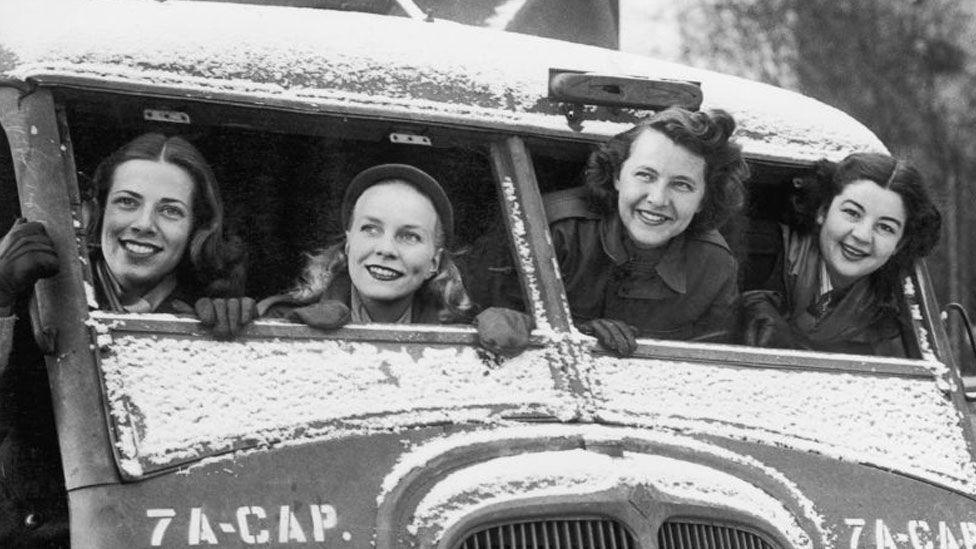 Four American Red Cross Clubmobile women in a captured German vehicle in France during World War II.  They were serving with the 36th Infantry Division. They are all looking out of the unglazed windshield of the vehicle and smiling broadly. Snow rests on the vehicle. From left to right Dorothy Boschen, Virginia Spetz, Jane Cook and Meredythe Gardiner.