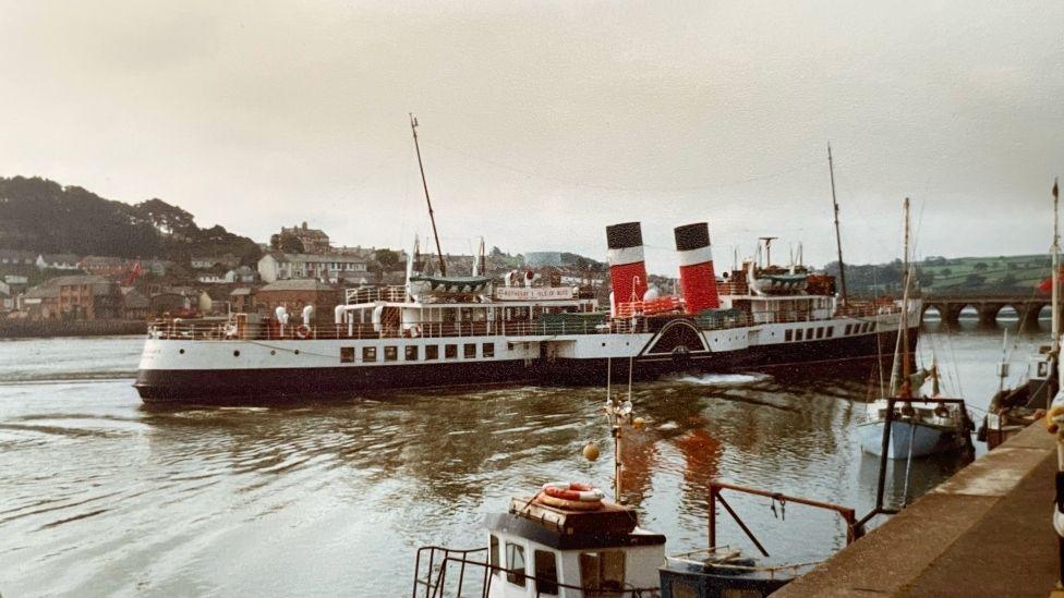 The Waverley paddle steamer in Bideford Harbour. The 240ft-long ship, operating since 1947, has an elongated hull; two black, white and bright red funnels, polished wood interiors and outside railings, with white and black striping along the sides under its open-air decks. 