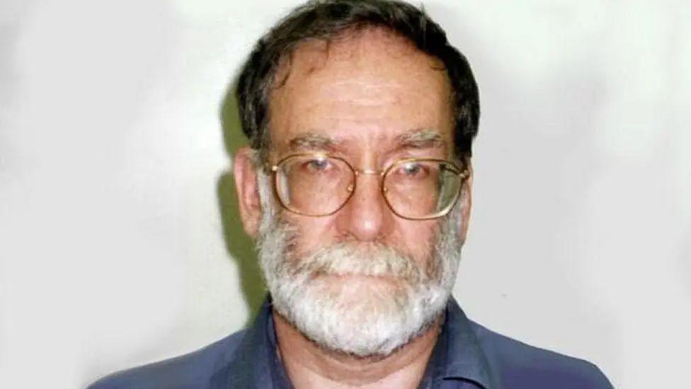 A mugshot of serial killer Harold Shipman, who has a white beard, wearing a blue shirt and wearing rectangular glasses.