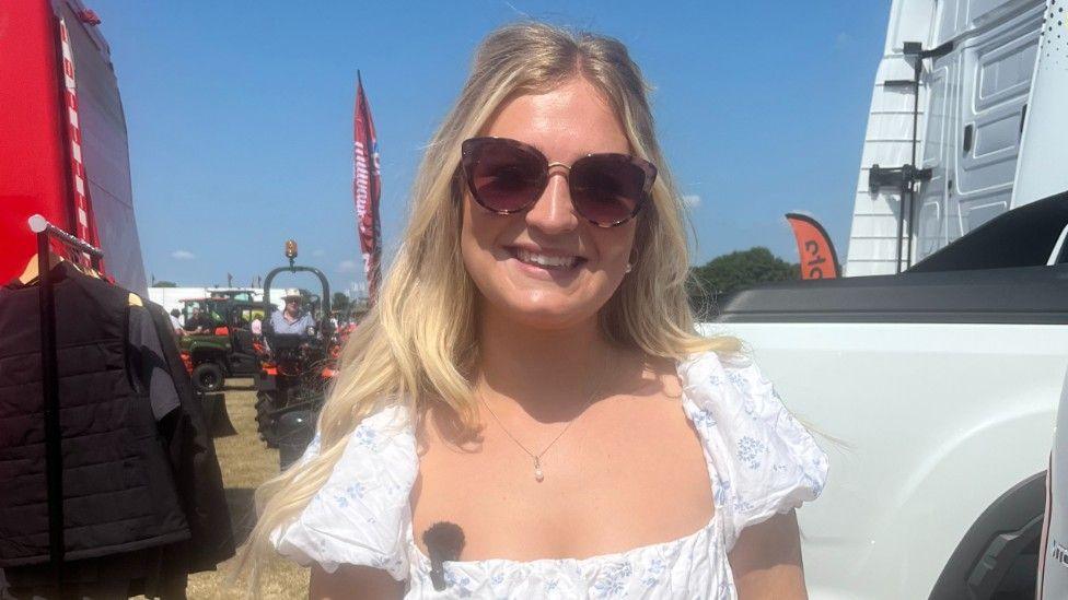 A woman with long blonde hair. She is wearing sunglasses, a white summer dress  and a necklace.