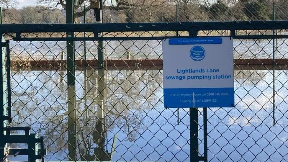 A metal fence with a Thames Water sign reading 'Lightlands Lane sewage pumping station' with a large reservoir of water behind it