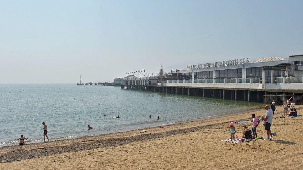 Small groups of people on the sand on a sunny day at Clacton beach. A long pier is in the background, stretching out to sea.