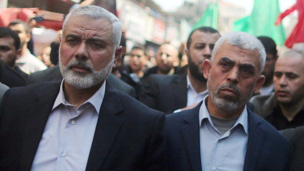 Ismail Haniyeh (left) and Yahya Sinwar (right) wear open-necked shirts with a dark suit jacket amongst a crowd of people on a busy street.