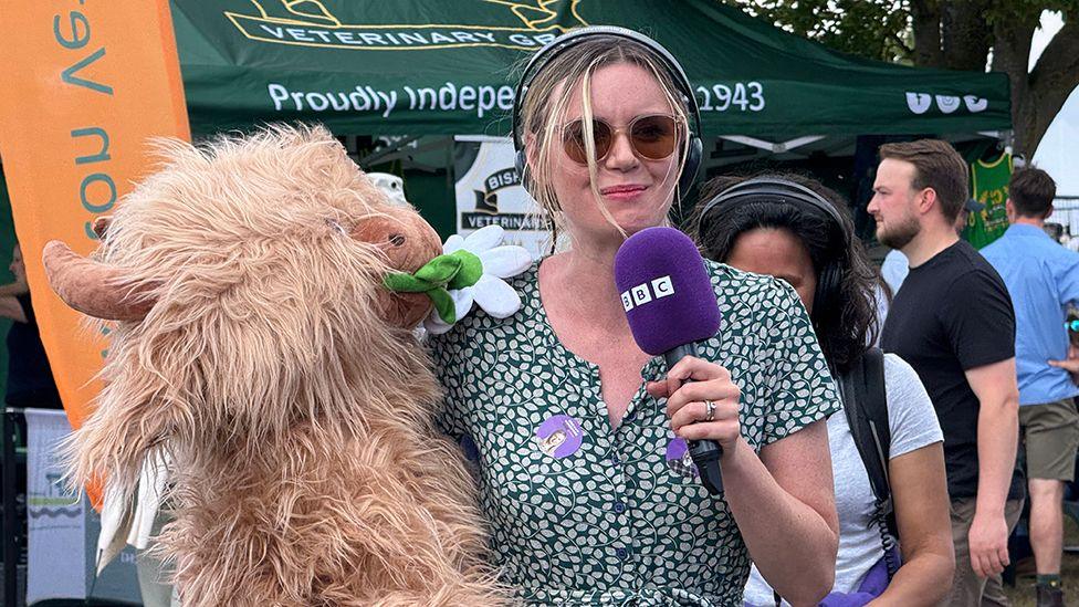 Radio Humberside presenter Carla Greene in green and white patterned dress and wearing sunglasses, headphones and holding a microphone holds a toy Highland Cow which she won from a stall