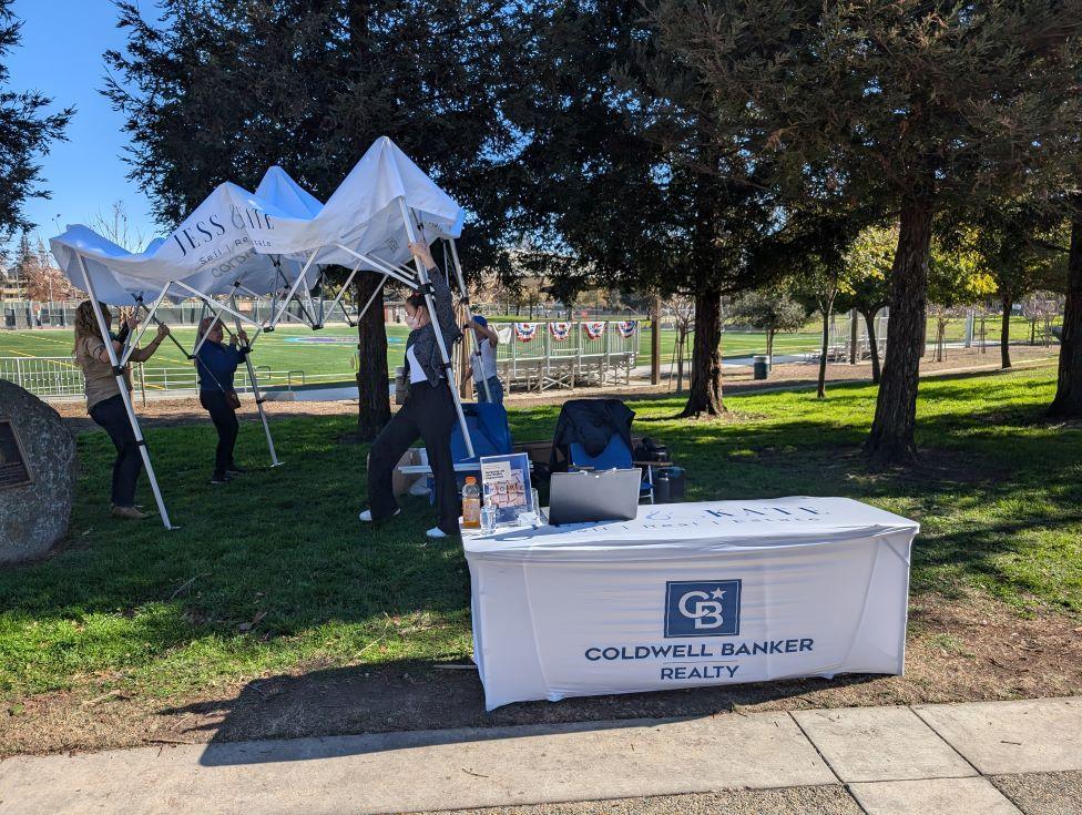 People take down a canopy behind a table offering realtor services, located in front of a park