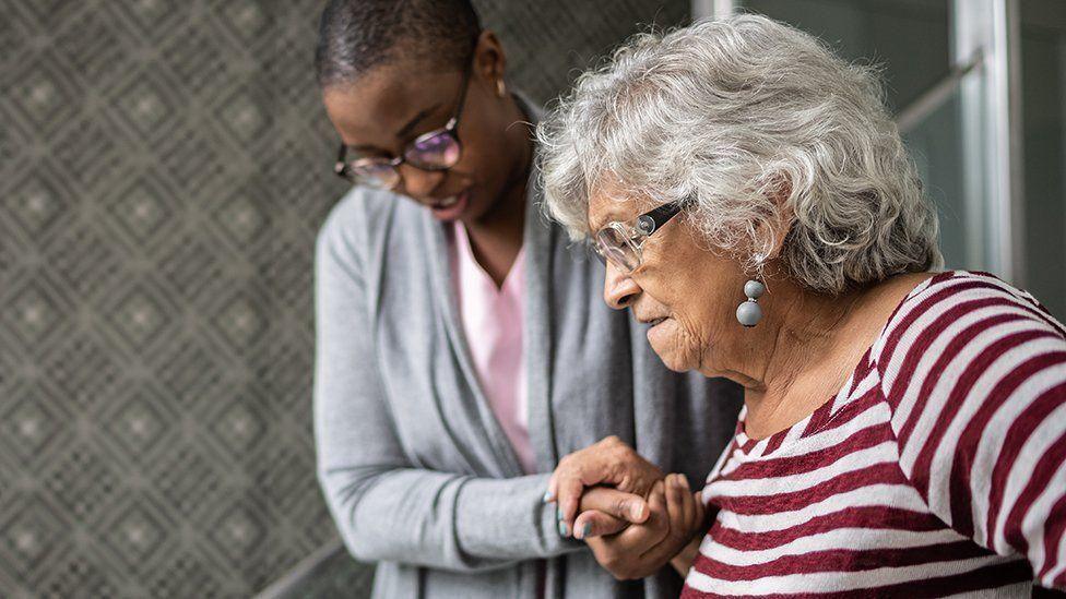 An elderly woman is helped to walk by a carer. The elderly woman, visible from the chest up, has grey curly hair and is wearing a red and white striped top. Her right hand is being held by a carer, with short black hair and glasses, wearing a grey cardigan and pink top. 