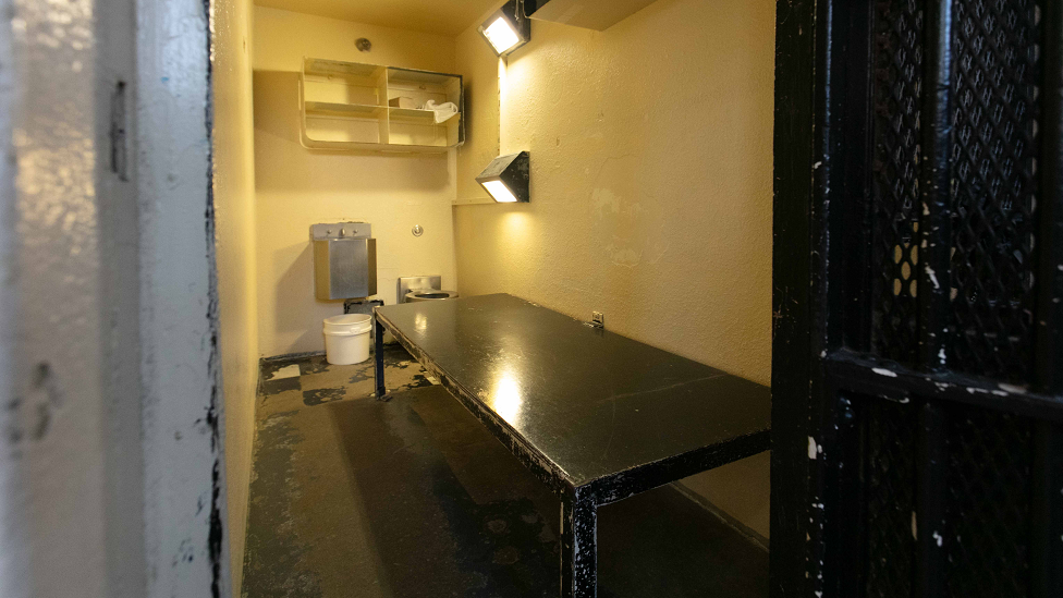 A photo of one of the cells, with a black table, a few lights, shelves, and a toilet