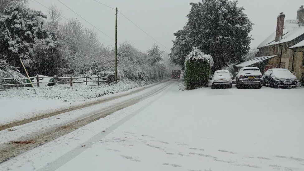 Snow covers road, cars and homes along a rural street.