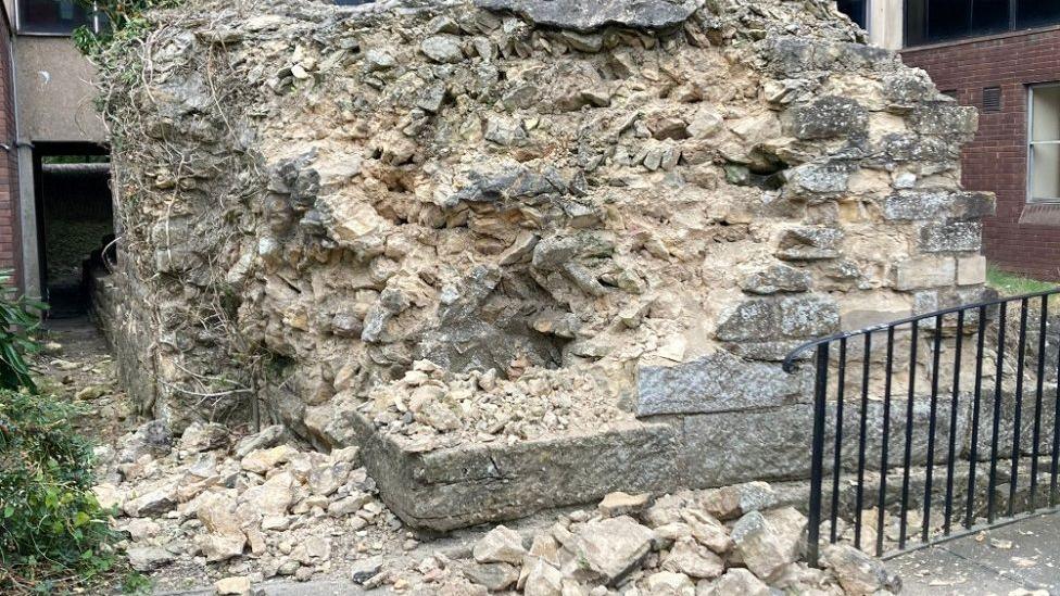 Light coloured bricks are seen damaged and in a pile of rubble, with a black railing and green bushes next to the wall