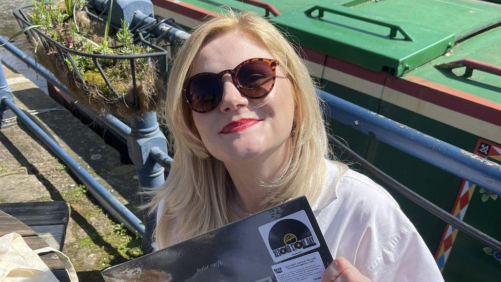 Lorna Paterson is holding a Taylor Swift vinyl, in a white t-shirt in front of a boat. She's wearing brown sunglasses as she smiles to the camera